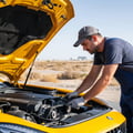 Inspector leaning over fender checking AC compressor belt and clutch, hood open, Dubai summer sun beating down, sweat on forehead