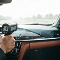 Inspector holding infrared thermometer aimed at dashboard AC vent, digital display showing 16 degrees, cold air ribbons visible, bright Dubai heat outside windshield