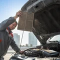 Inspector lifting dirty air filter out of filter box, holding it up to sunlight showing dust buildup, clean engine bay behind, outdoor Dubai