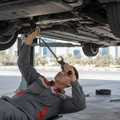 Inspector under car prying ball joint with bar checking for play, dust boot visible, flashlight in other hand, car on lift Dubai outdoor