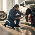 Inspector kneeling beside front wheel removed, examining brake caliper and rotor, Dubai parking lot, morning shadow under car