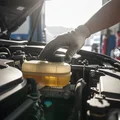 Inspector under hood pressing brake fluid reservoir cap, checking fluid level and color against MIN MAX marks, bright engine bay outdoor light