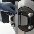 Close-up of gloved hand sliding brake pad out of caliper bracket, measuring remaining thickness with ruler, rotor surface behind, bright outdoor