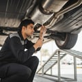 Inspector tapping catalytic converter heat shield with wrench, listening for rattle, car on ramps, bright outdoor light underneath