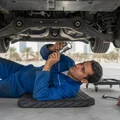 Inspector under front end checking control arm bushings for cracks, rubber split visible, bright flashlight beam, car on ramps outdoor Dubai