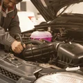 Inspector squeezing coolant hose while checking overflow tank level, pink fluid behind transparent wall, steam-free radiator cap area, Dubai afternoon heat