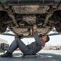Close-up of inspector's hand squeezing CV boot at front wheel, checking for grease leak or tear, wheel turned full lock, bright Dubai outdoor