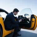 Inspector leaning into open driver door checking digital gauge cluster, finger scrolling through trip computer, bright morning light on display