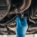 Inspector following exhaust pipe from manifold to tail, crawling under car with flashlight checking each hanger mount, outdoor Dubai daylight