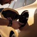 Inspector opening fuel filler cap, sniffing for vapor, capless funnel neck visible inside, car side panel in bright Dubai afternoon sun