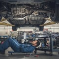 Inspector pointing flashlight at fuel filter bracket underneath car, checking for rust or line corrosion, ground dry below, bright Dubai outdoor light from sides