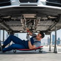Inspector under car on creeper following fuel line from tank forward, flashlight checking fittings for drips, car on ramps outdoor Dubai daylight