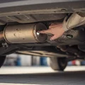 Inspector under rear of car pointing at muffler weld seam, checking for rust holes, exhaust pipe running forward, bright Dubai outdoor daylight