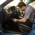 Inspector kneeling at open driver door plugging OBD scanner cable into port under steering column, blue LED on connector glowing, bright Dubai daylight flooding cabin