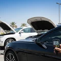 Inspector holding paint thickness gauge on SUV hood with diagnostic tablet on fender, bright outdoor Dubai sunlight
