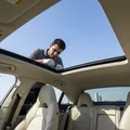 Interior shot looking up through open panoramic sunroof, inspector leaning over roof checking glass edges, blue Dubai sky above