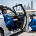 Inspector pulling parking brake lever inside car while assistant checks rear wheel won't spin, view from open rear door, Dubai outdoor daylight
