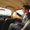 Inspector sitting in rear seat holding thermometer at back AC vent, checking rear passenger cooling, bright Dubai afternoon light through rear window