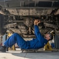 Inspector at rear of lifted car spinning rear wheel checking differential noise, hand on axle housing feeling vibration, bright Dubai afternoon sun