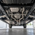 Rear frame rails and spare tire well visible from underneath, inspector using flashlight checking for accident damage, car lifted in bright Dubai parking