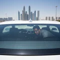 Rear of SUV, inspector crouching to check rear windshield from outside, defroster lines visible through glass, bright Dubai parking