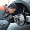 Inspector at front wheel checking regenerative brake wear pattern on rotor, less wear than conventional, electric motor housing visible behind, bright outdoor