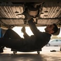 Close-up of inspector wiggling rubber exhaust hanger checking elasticity, pipe joint visible above, bright outdoor undercarriage light