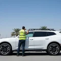 Inspector stepping on running board testing weight, then checking roof rail mounting bolts, SUV side view, bright Dubai outdoor palm trees