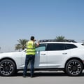 Inspector stepping on running board testing weight, then checking roof rail mounting bolts, SUV side view, bright Dubai outdoor palm trees