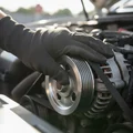 Close-up of inspector's gloved hand pressing serpentine belt checking tension, alternator pulley visible behind, bright outdoor light filling engine bay