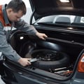 Inspector lifting trunk floor mat revealing spare tire, checking pressure with gauge, jack and tools beside it, bright trunk interior light
