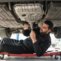 Inspector lying on creeper under car tapping transmission pan with wrench listening for sludge, oil drain plug visible, outdoor bright Dubai light