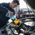 Inspector topping up blue washer fluid from bottle, funnel in reservoir neck, power steering cap open beside it for check, bright outdoor engine bay sun