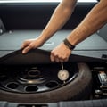Inspector spinning front wheel listening to bearing, hand on strut feeling for vibration, car on jack stands, bright outdoor Dubai parking