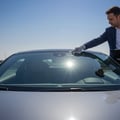 Inspector standing outside car pointing at small chip on windshield corner, Dubai parking lot, morning sun reflecting palm trees on glass