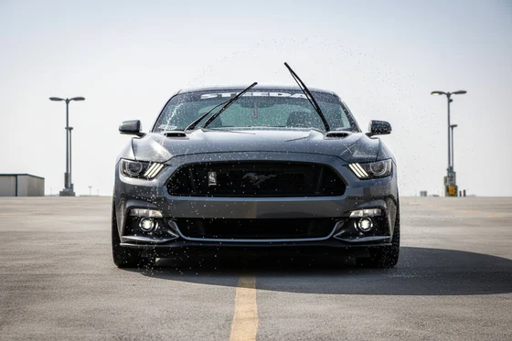 Inspector activating windshield wipers from inside, washer fluid spraying on glass, wipers sweeping clean, view from outside, bright Dubai outdoor